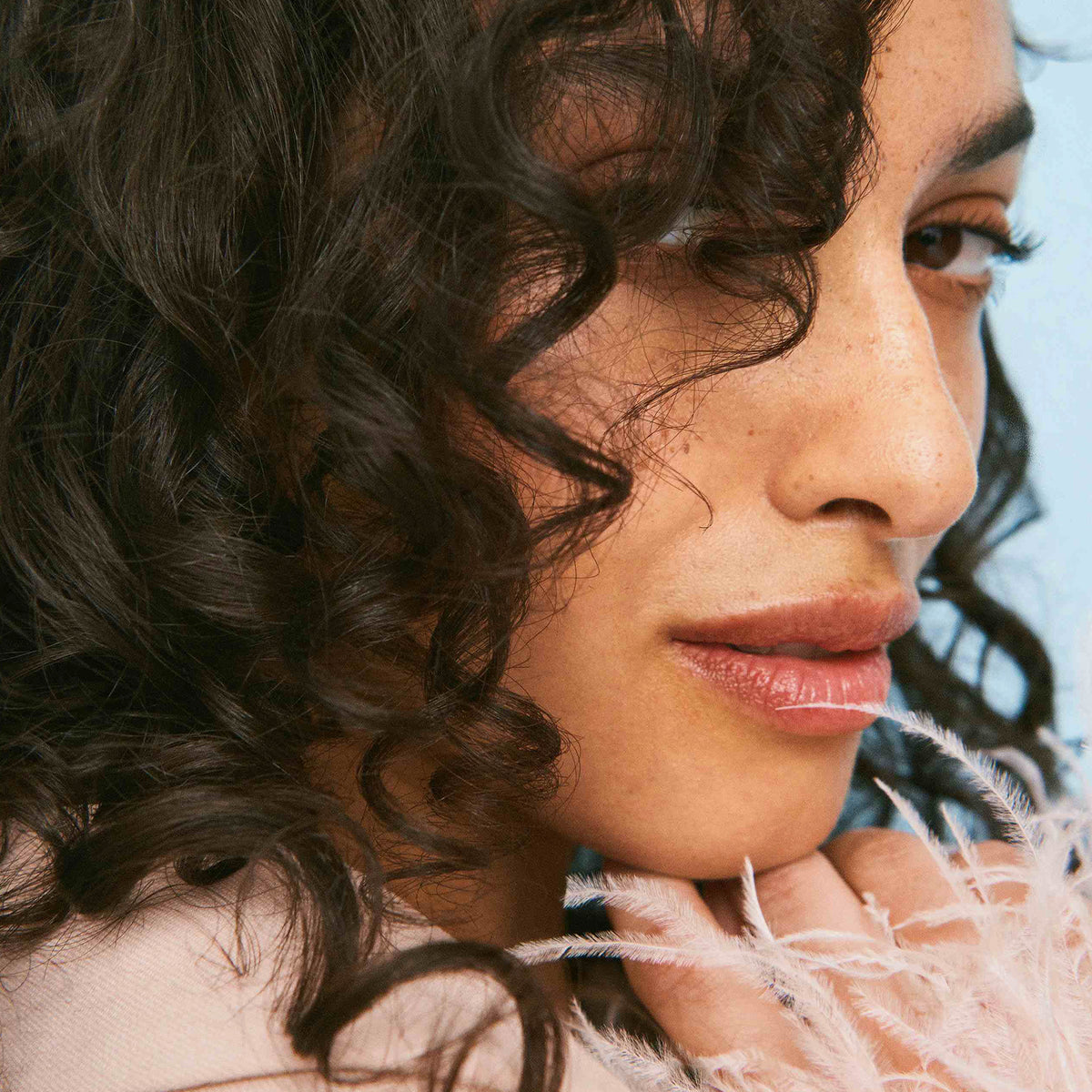 Eine Frau mit lockigem Haar und natürlichem Make-up blickt zur Seite und präsentiert Ergebnis des Coils & Curls Weightless Curl Definer von Maria Nila.