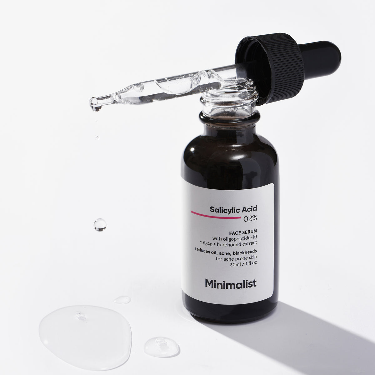 Eine Flasche Minimalist Salicylic Acid 02 % Face Serum mit einer Pipette und Serumtropfen auf einer weißen Oberfläche.