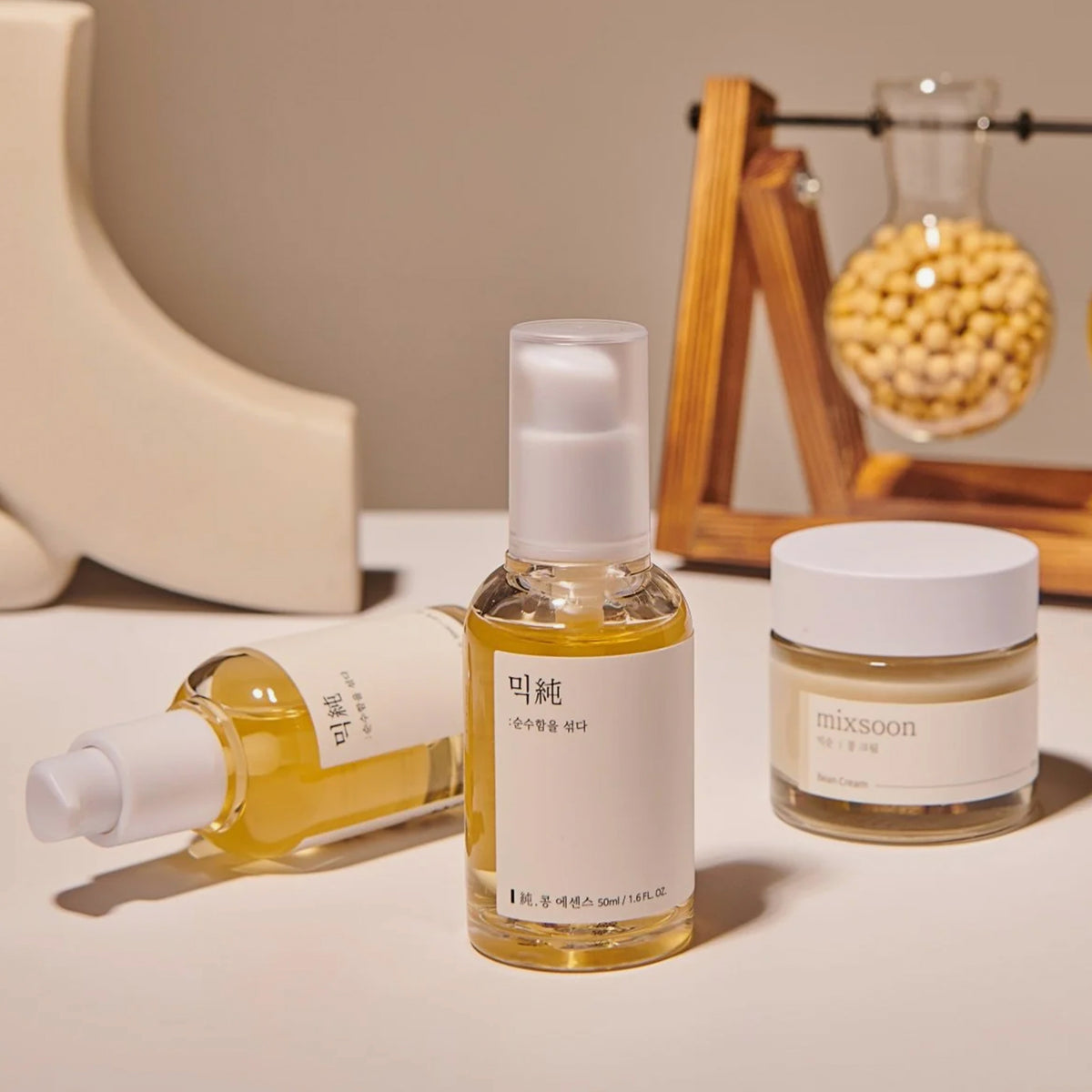 Drei Mixsoon Bean Hautpflegeprodukte - mit minimalistischen Etiketten auf heller Oberfläche; im Hintergrund Sojabohnen.