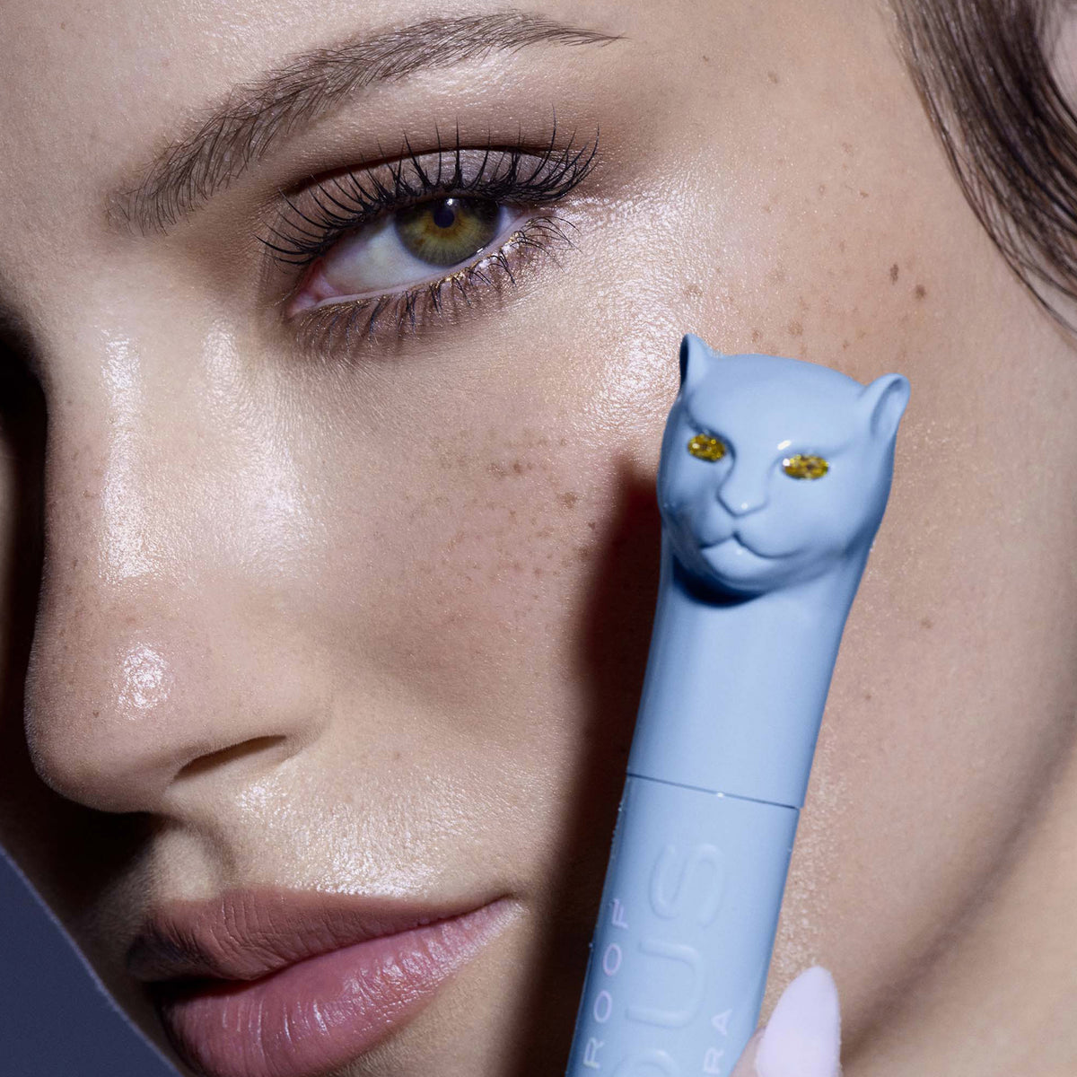 Eine Frau hält Vicious Mascara Waterproof mit einer katzenförmigen Kappe vor ihr Gesicht und betont so ihre langen Wimpern.