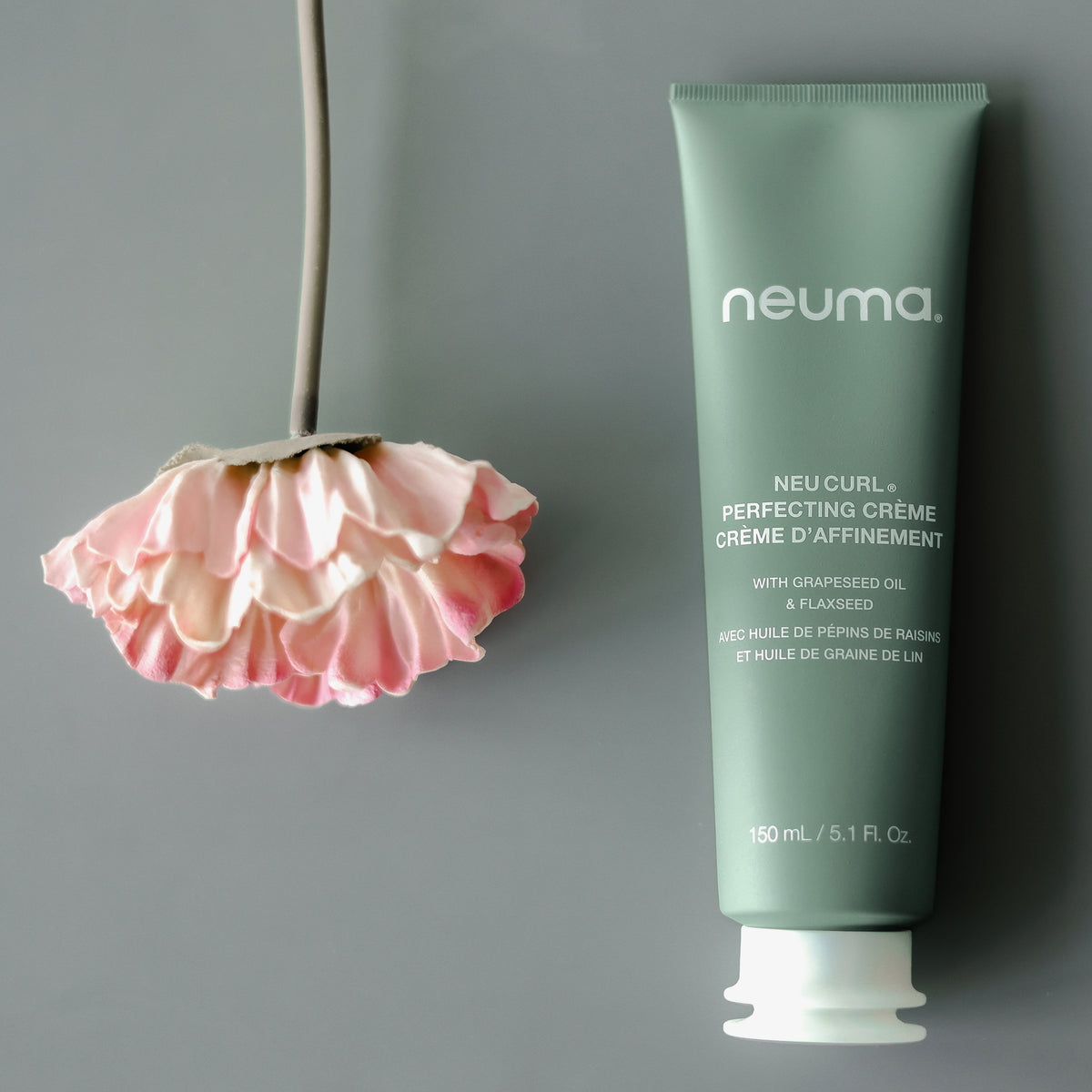 Eine rosa Blume hängt kopfüber neben einer Tube Neuma NEU CURL™ PERFECTING CRÈME auf einem grauen Hintergrund.