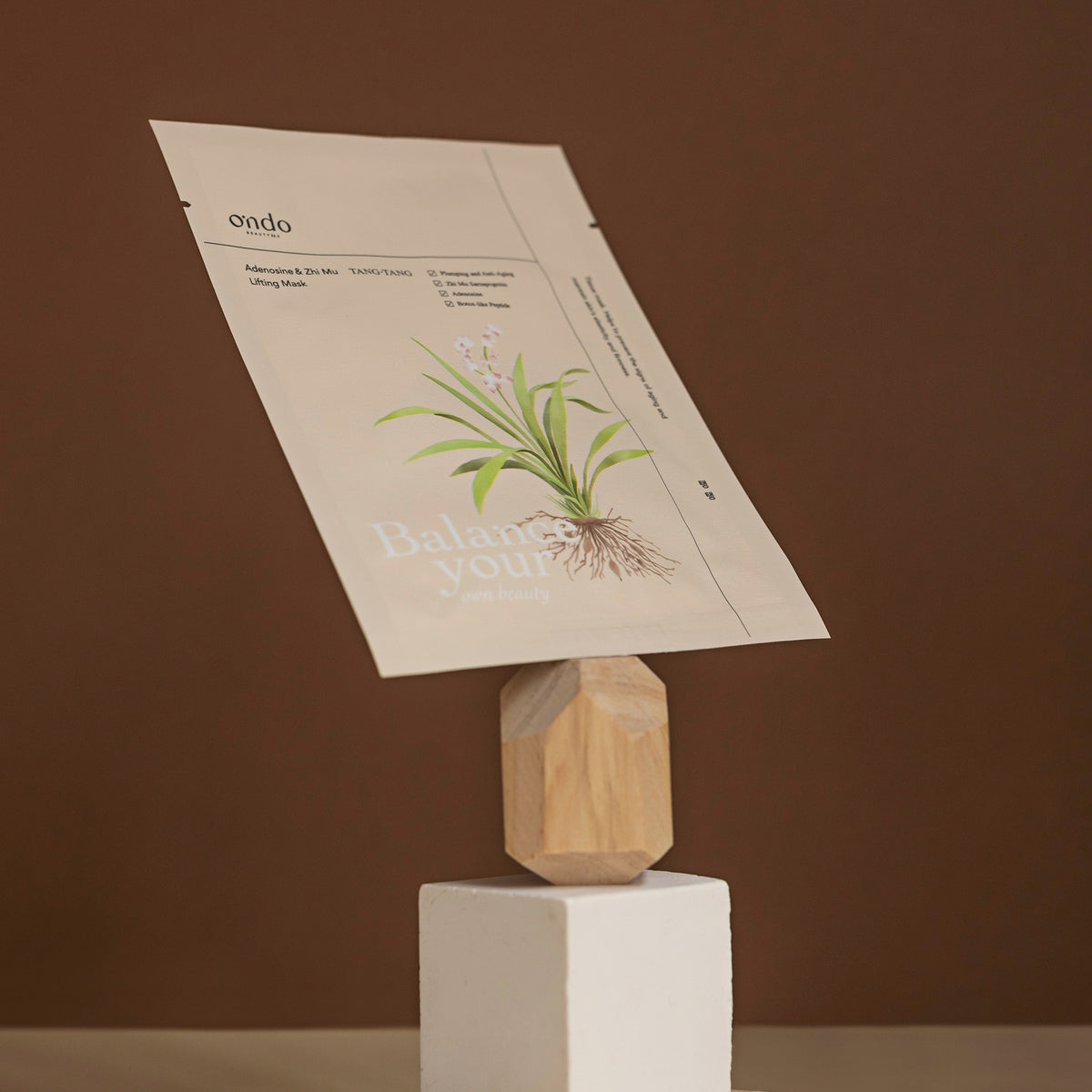 Die Adenosine & Zhi Mu Lifting Mask Tang-Tang in einer beigen Packung balanciert auf einem geometrischen Holzständer vor braunen Hintergrund.