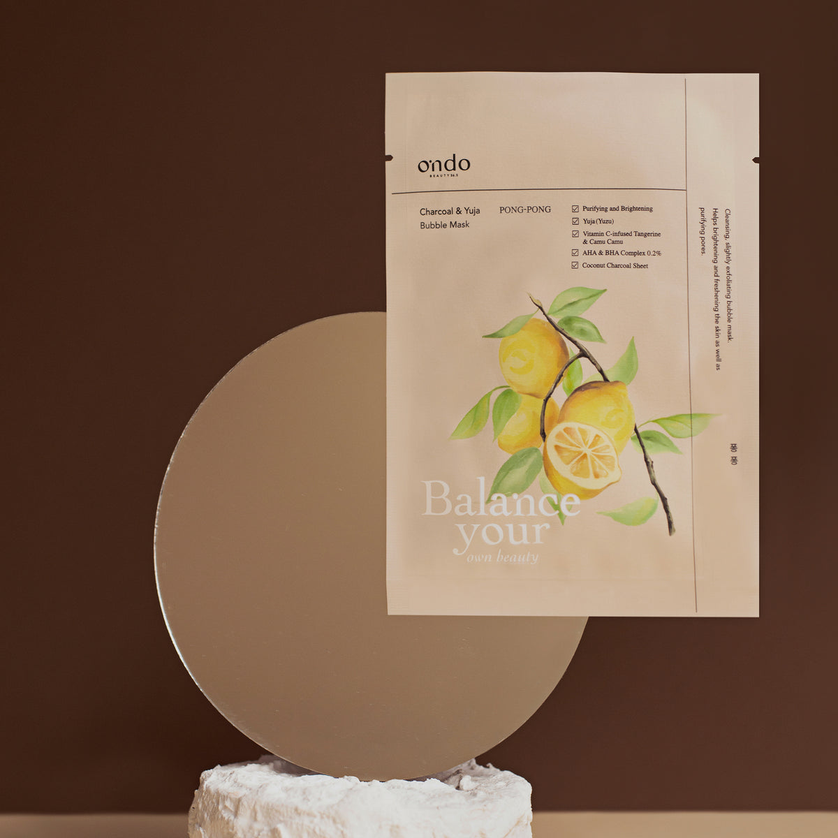 Das Pong-Pong-Paket mit der Charcoal & Yuja Bubble Mask mit Illustrationen wird vor einem braunen Hintergrund mit einem runden Spiegel präsentiert.