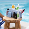 Die PURISH Summer Bag 2025 – Deluxe Routine liegt auf einem Holzhocker neben dem Pool und bietet eine Sammlung wichtiger Hautpflegeprodukte in einer stilvollen blauen Kulturtasche.