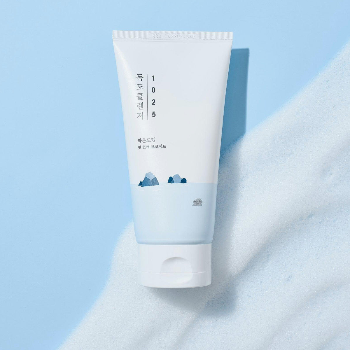 Weiße Tube des Round Lab 1025 Dokdo Cleansers mit blauen Akzenten und Inselmotiv, auf hellem Untergrund mit aufgeschäumtem Produkt abgebildet.