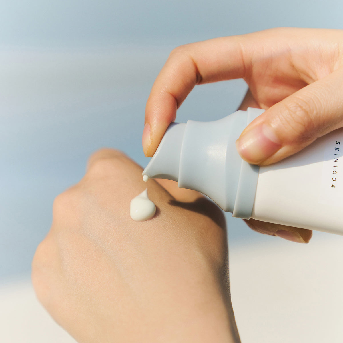 Vor einem sanften blauen Hintergrund pumpt eine Hand Madagascar Centella Hyalu-Cica Water-Fit Sun Serum auf den Handrücken der anderen Hand.