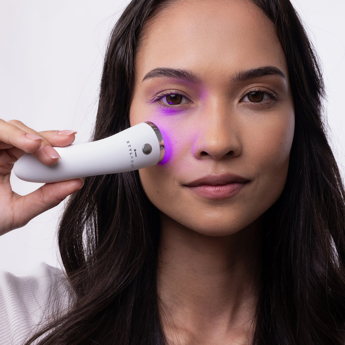 Eine Frau benutzt den STYLPRO Brightspot Anti-Blemish LED-Stab, ein weißes Handgerät mit lila Licht, auf ihrem Gesicht, während sie in die Kamera schaut.