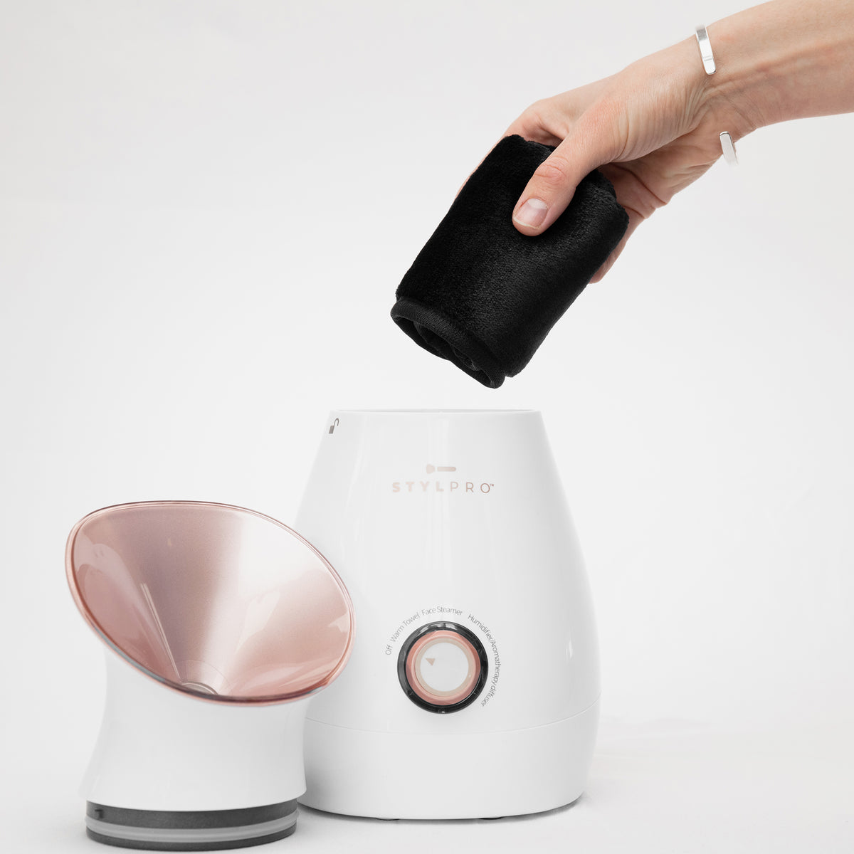 Eine Hand hält ein schwarzes Tuch über den Facial Steamer mit weißem Gehäuse und rosa Düse vor einem schlichten Hintergrund.