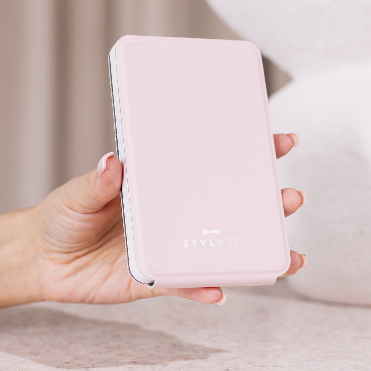 Eine Hand hält einen geschlossenen, rosafarbenen STYLPRO LED-Minispiegel mit abgerundeten Kanten auf einer hellen, strukturierten Oberfläche.