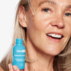 Eine Frau lächelt, während sie Tropfen Hyaluronsäure-Vitamin-C-Serum aus einer blauen Flasche aufträgt.