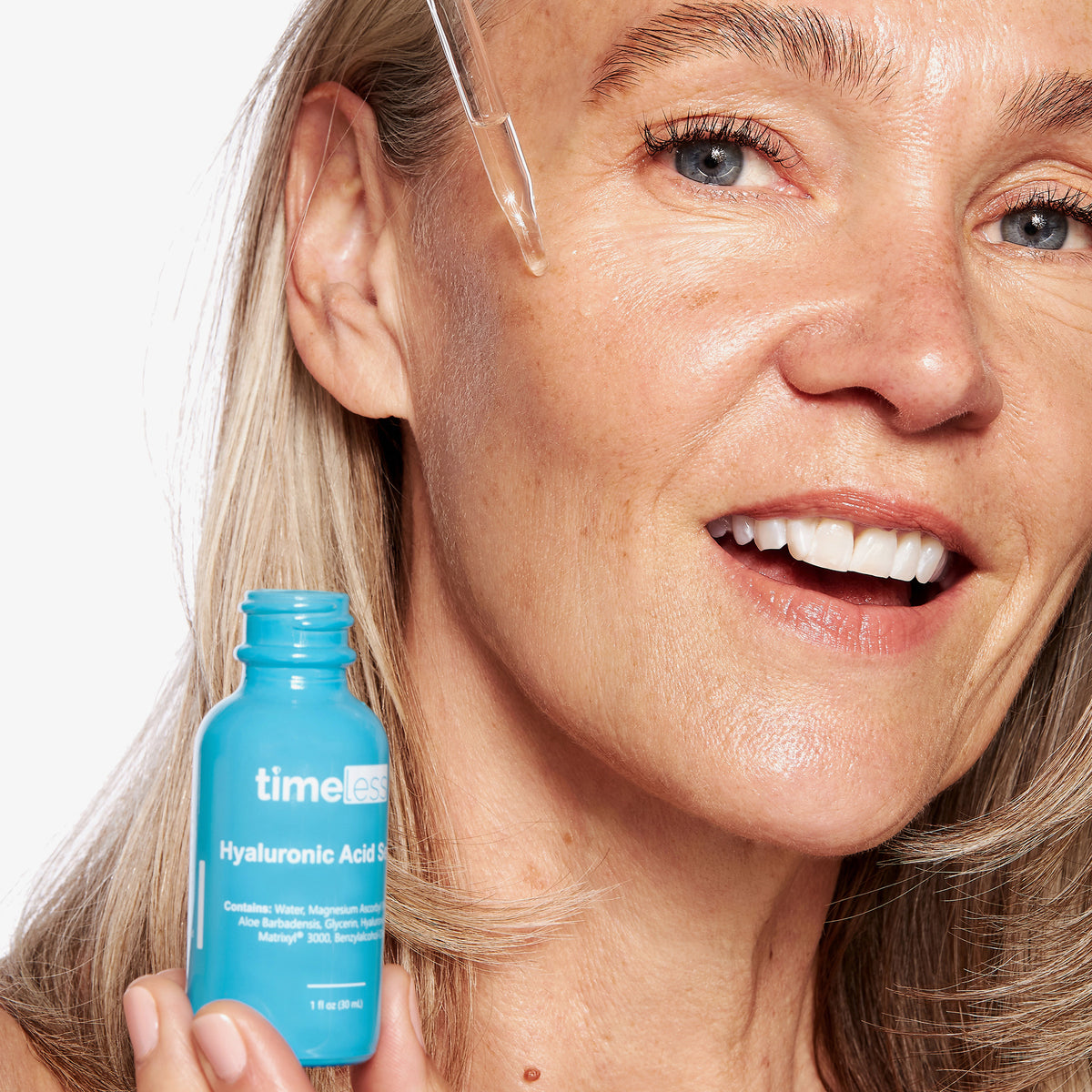 Eine Frau lächelt, während sie Tropfen Hyaluronsäure-Vitamin-C-Serum aus einer blauen Flasche aufträgt.