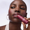 Nahaufnahme einer Frau die den Blushin' Charm Multi Stick 030 auf den Wangen und Lippen trägt und in die Kamera hält.