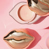 KISS & GLOW Shimmer Lip Balm C01 in lippenförmigem Behälter mit goldenem Deckel auf rosa Oberfläche.