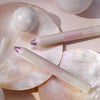 Zwei lilane PEARLFECTION Eyeshadow Sticks von Catrice auf changierenden Muscheln mit dekorativen Perlen.