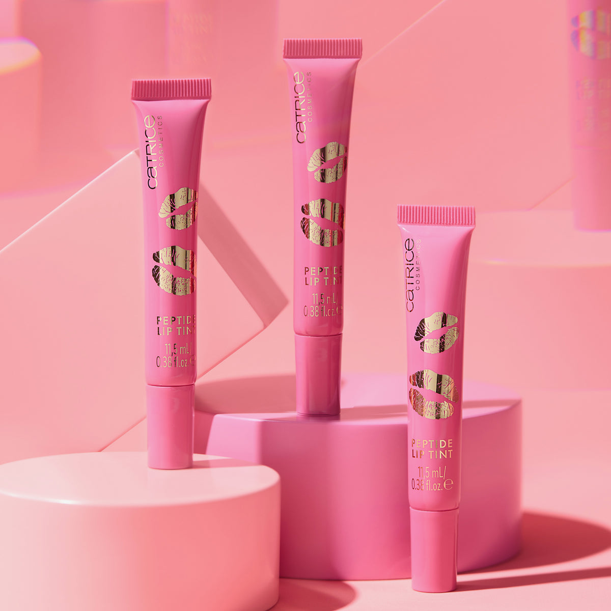 Drei KISS & GLOW Peptide Lip Tint C01-Tuben auf rosa geometrischen Plattformen vor einem rosa Hintergrund.