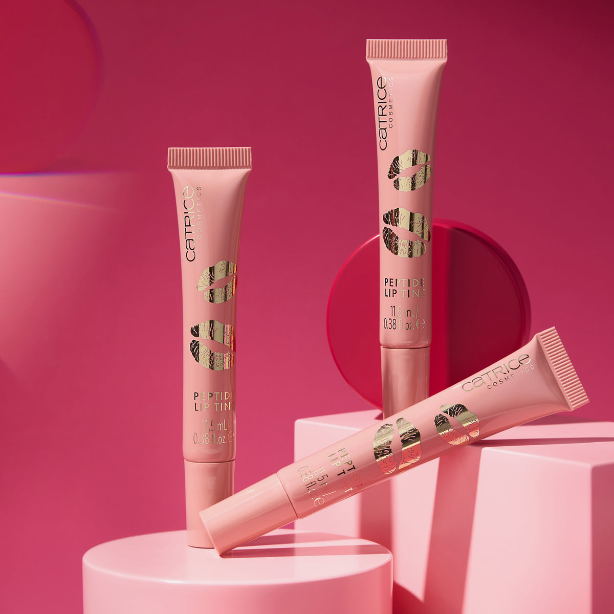 Drei nudefarbene Tuben KISS & GLOW Peptide Lip Tint C02 vor einem leuchtend rosa Hintergrund mit geometrischen Formen.