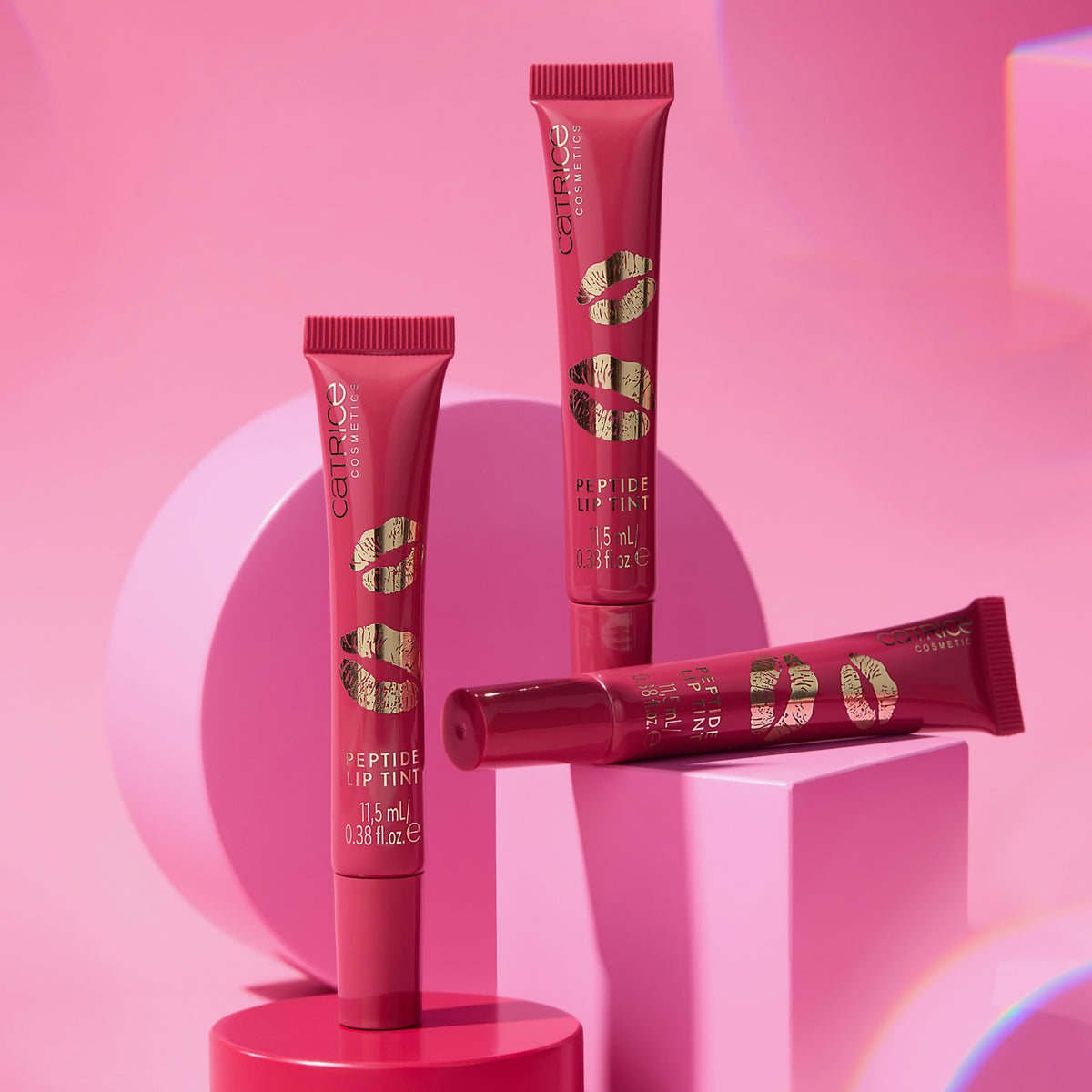 Drei Tuben KISS & GLOW Peptide Lip Tint C03 geometrischen Plattformen vor rosa Hintergrund.