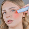 Eine Frau trägt den Blush Stick – 2-in-1 Creme Rouge mit Pinsel Love auf ihre Wange auf und blickt in die Kamera.