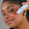 Eine Frau trägt den Blush Stick – 2-in-1 Creme Rouge mit Pinsel Scarlet auf ihre Wange auf und blickt in die Kamera.