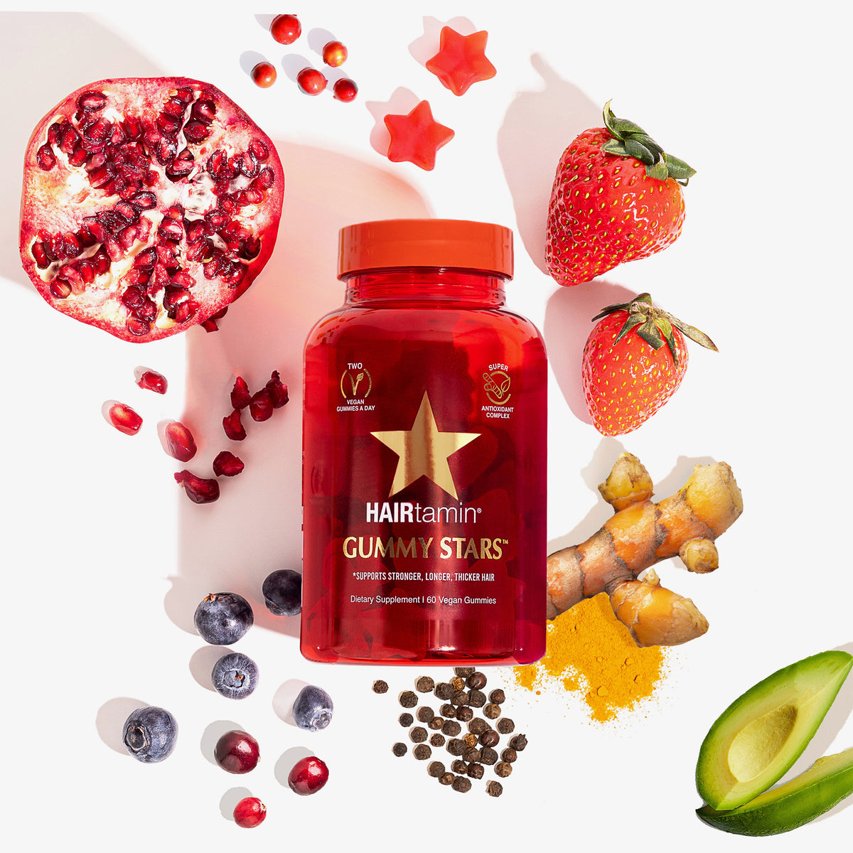Rote Flasche HAIRtamin Gummy Stars umgeben von Früchten, Gewürzen und Avocadoscheiben auf weißem Hintergrund.