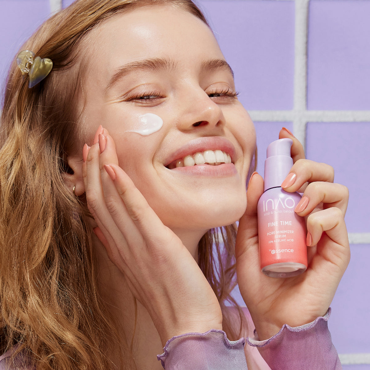 Eine lächelnde Frau trägt Gesichtscreme auf und hält eine rosa Flasche Fine Time Pore Minimizer Serum in der Hand.