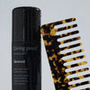 Eine Flasche Living Proof Style Lab Blowout Spray in Schwarz steht neben einem braunen Schildpatt-Breitzahnkamm auf einem hellen Hintergrund.