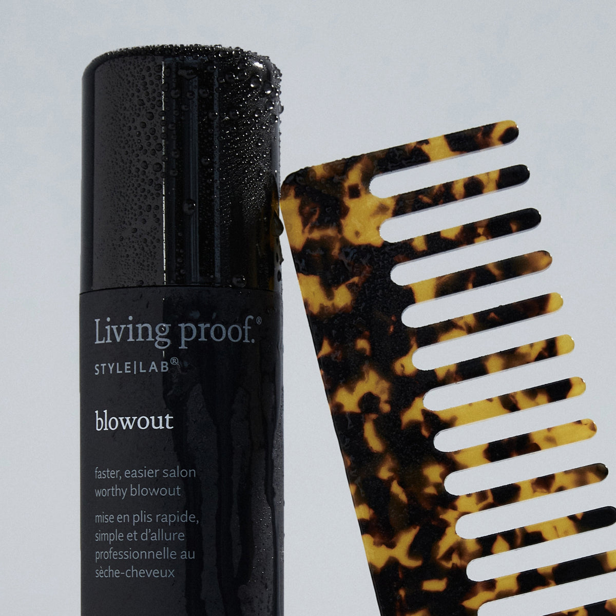 Eine Flasche Living Proof Style Lab Blowout Spray in Schwarz steht neben einem braunen Schildpatt-Breitzahnkamm auf einem hellen Hintergrund.