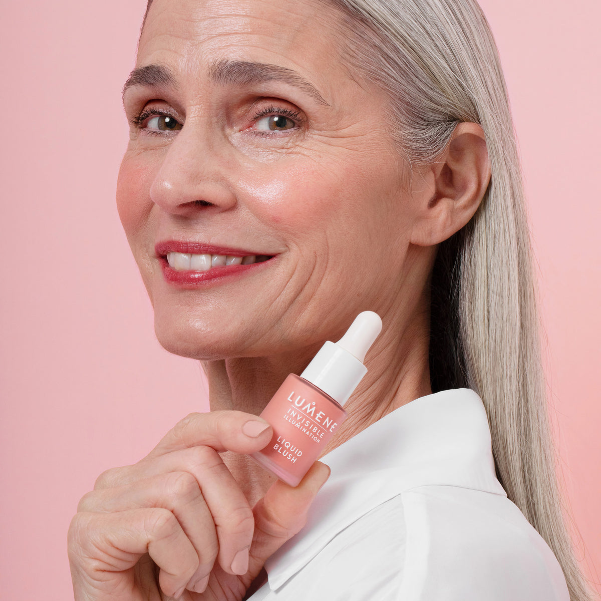 Eine Frau mit strahlendem hellen Hautton und grauem Haar hält eine Flasche Lumene INVISIBLE ILLUMINATION Liquid Blush Pink Blossom an ihr Gesicht.