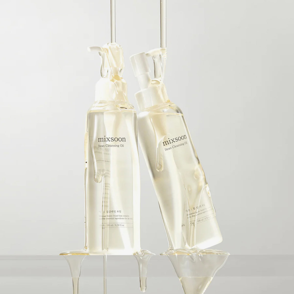 Zwei durchsichtige Flaschen mit Mixsoon Bean Cleansing Oil unter fließendem Öl , ausgestellt vor einem hellen Hintergrund.