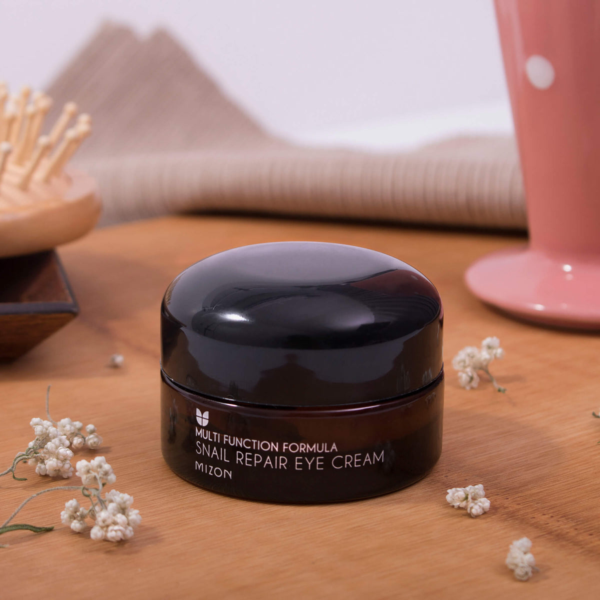 Ein Tiegel  Snail Repair Eye Cream steht auf einer Holzoberfläche, im Hintergrund sind Blumen und Dekoartikel zu sehen.