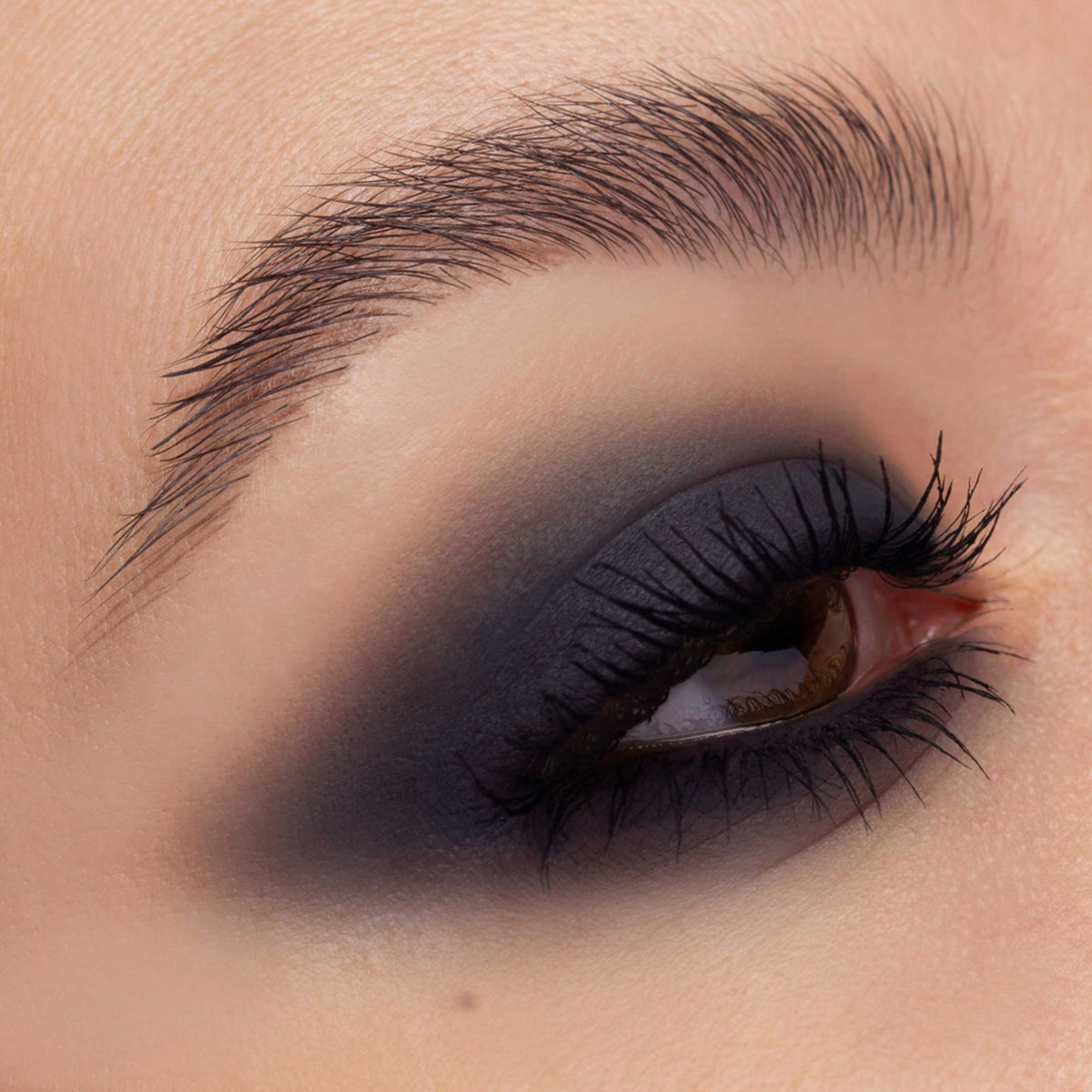 Nahaufnahme eines Auges mit dramatischem Smokey-Eye-Look in tiefem Anthrazit, weich verblendet bis über die Lidfalte hinaus. Die Wimpern sind lang und geschwungen.