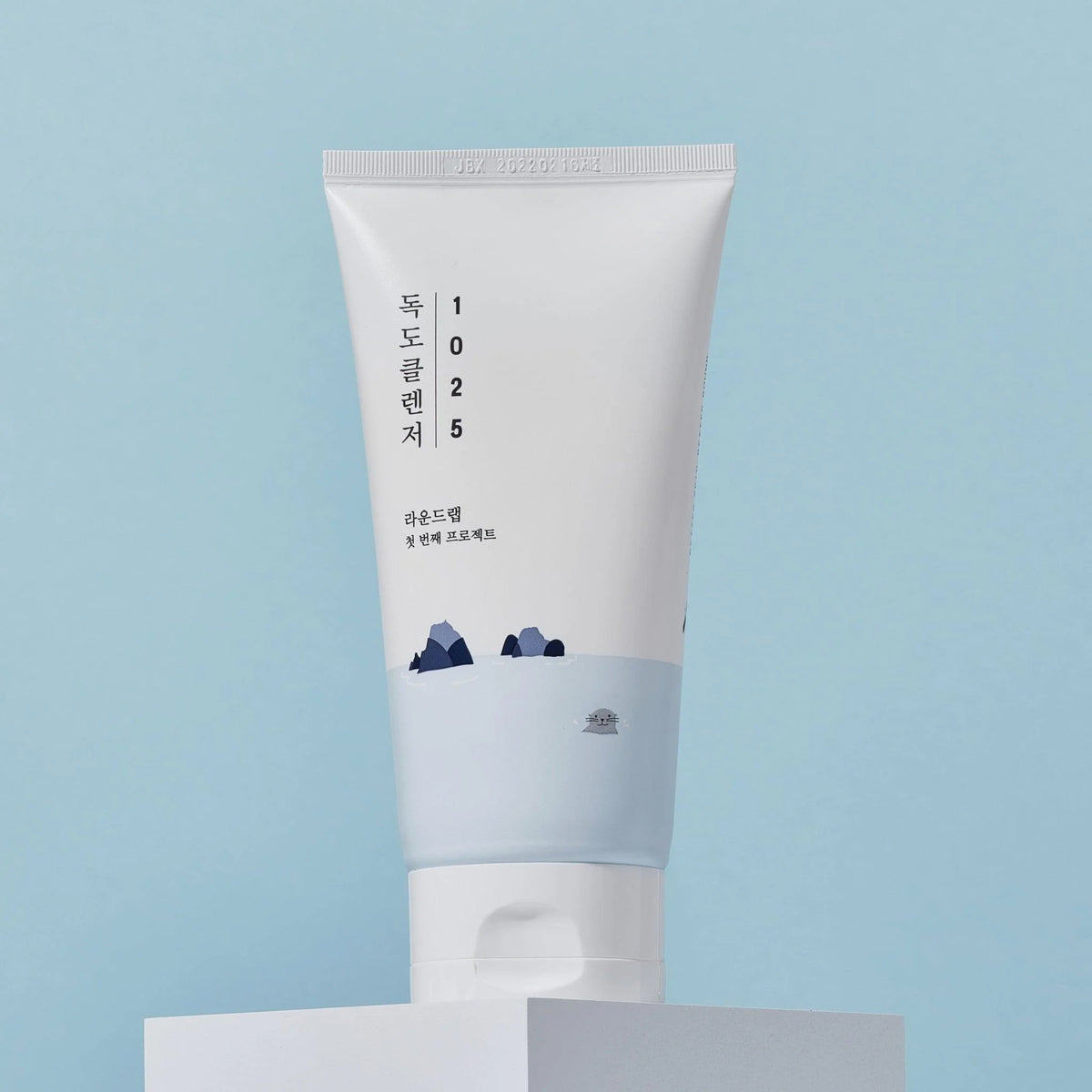 Eine Tube 1025 Dokdo Cleanser mit minimalistischem Design in Weiß und Blau steht auf einem weißen Block vor blauem Hintergrund.