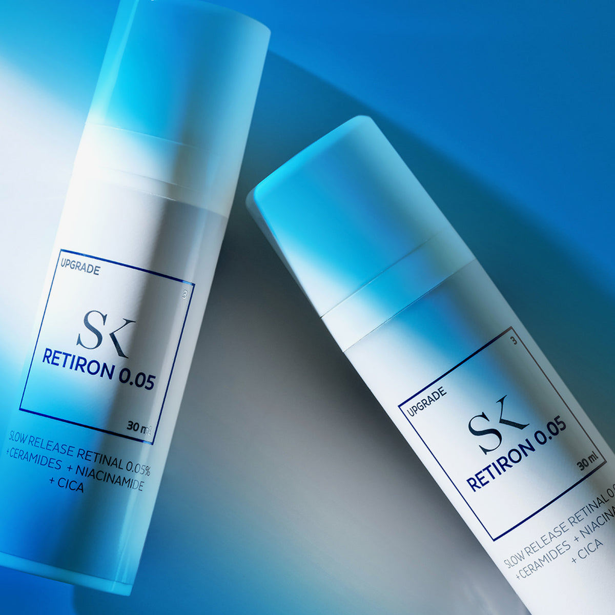 Zwei blau-weiße Retiron 0,05-Hautpflegeflaschen liegen auf einer reflektierenden Oberfläche mit einem blauen Farbverlaufshintergrund.