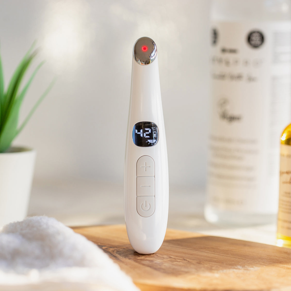 Elektronisches Beauty-Tool mit Display und Bedientasten, steht aufrecht auf Holzunterlage. Im Hintergrund Pflegeprodukte und Pflanze.