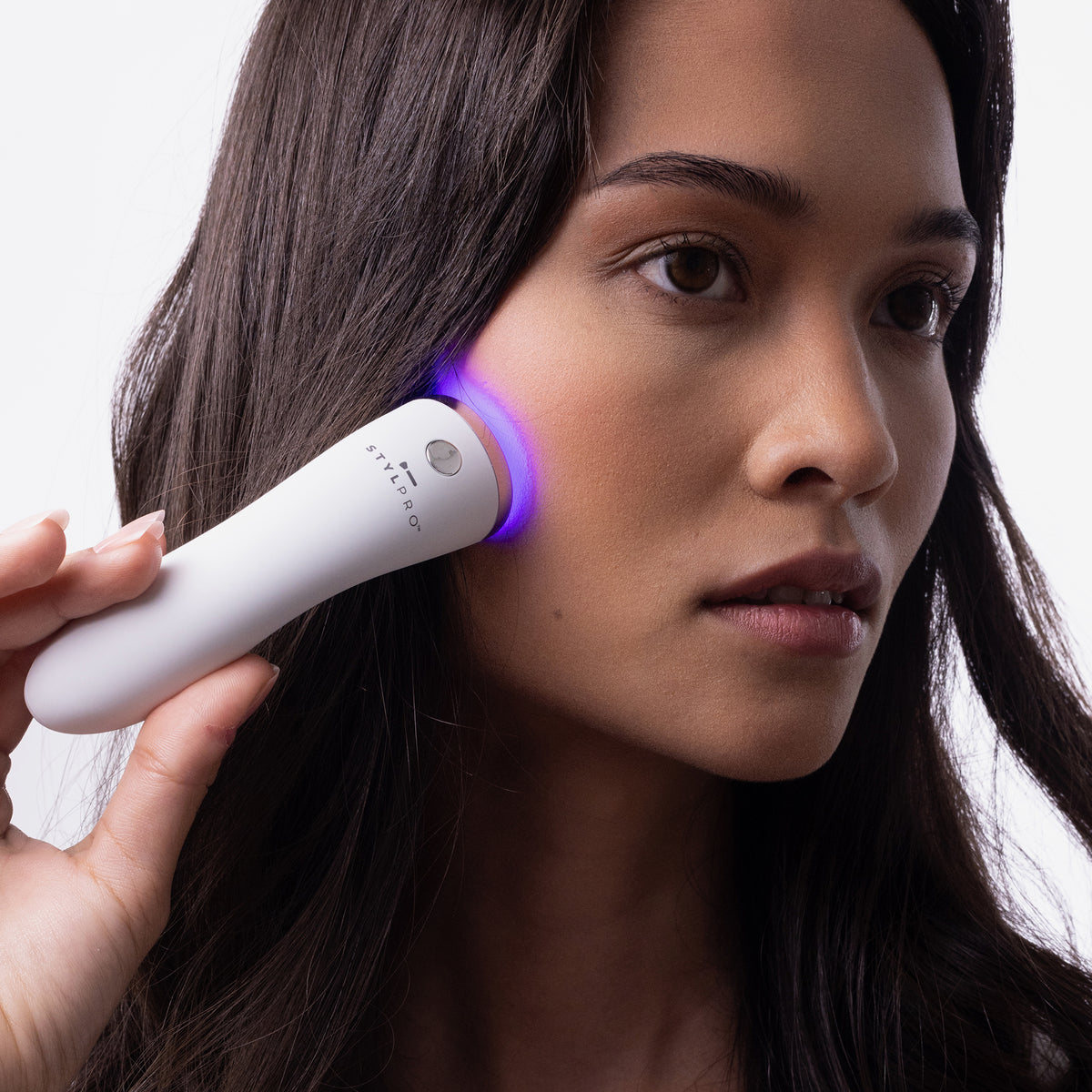 Eine Frau verwendet den STYLPRO Brightspot Anti-Blemish LED-Stab mit blauem Licht auf ihrer Wange vor einem neutralen Hintergrund.