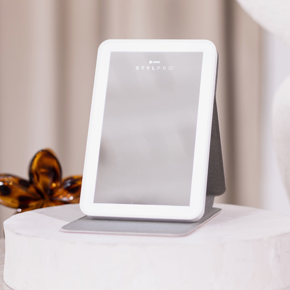 Der STYLPRO LED Mini Mirror ist ein rechteckiger Schminkspiegel mit weißem Rahmen, der auf jeder runden Oberfläche stehen kann.