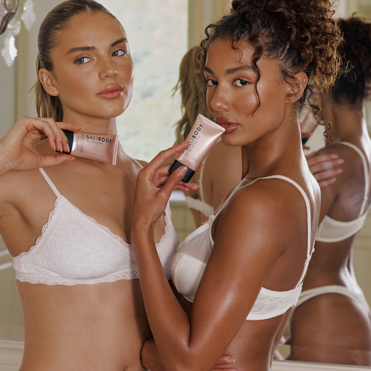 Zwei gebräunte Frauen in Dessous stehen vor einem Spiegel mit BB Creams in der Hand.