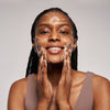 Lächelnde Frau wäscht ihr Gesicht mit dem AHA/PHA + Organic Aloe Vera Cleanser Gel vor einem schlichten Hintergrund.