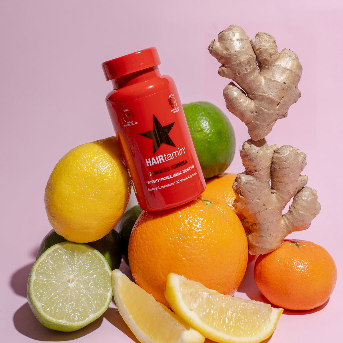 Eine Flasche HAIRtamin Advanced Formula 1 Month in Rot, auf rosa Hintergrund, umgeben von Zitrusfrüchten und Ingwerwurzel.