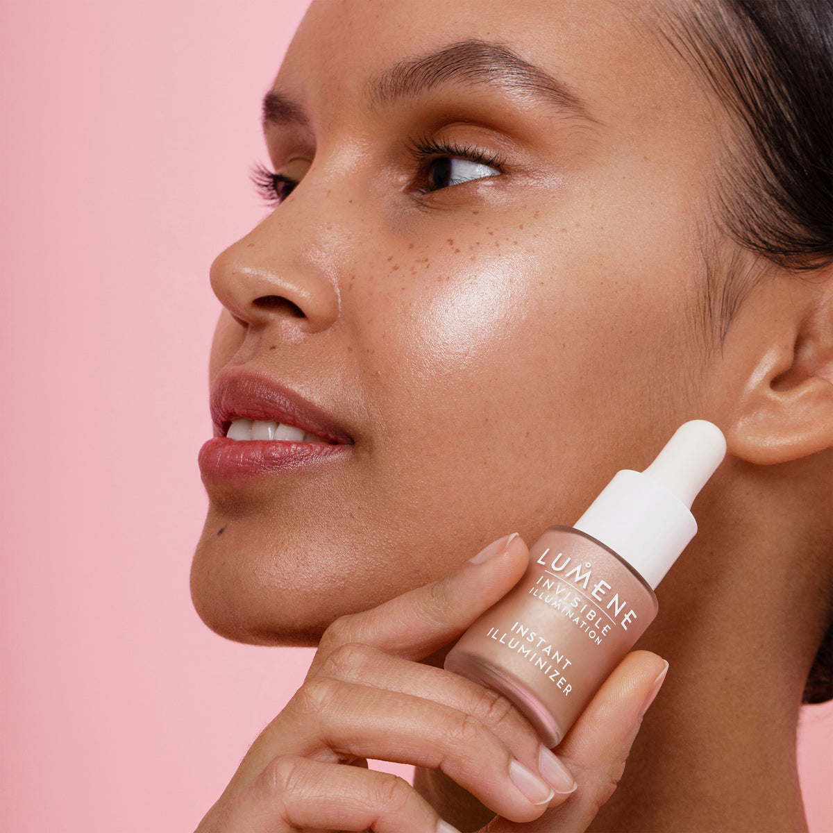 Eine Frau mit strahlender Haut hält eine Flasche Lumene INVISIBLE ILLUMINATION Illuminizer in der Nähe ihres Gesichts vor einem rosa Hintergrund.