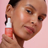 Eine Frau mit strahlendem mittlerem Hautton hält eine Flasche Lumene INVISIBLE ILLUMINATION Liquid Blush Bright Blossom an ihr Gesicht.