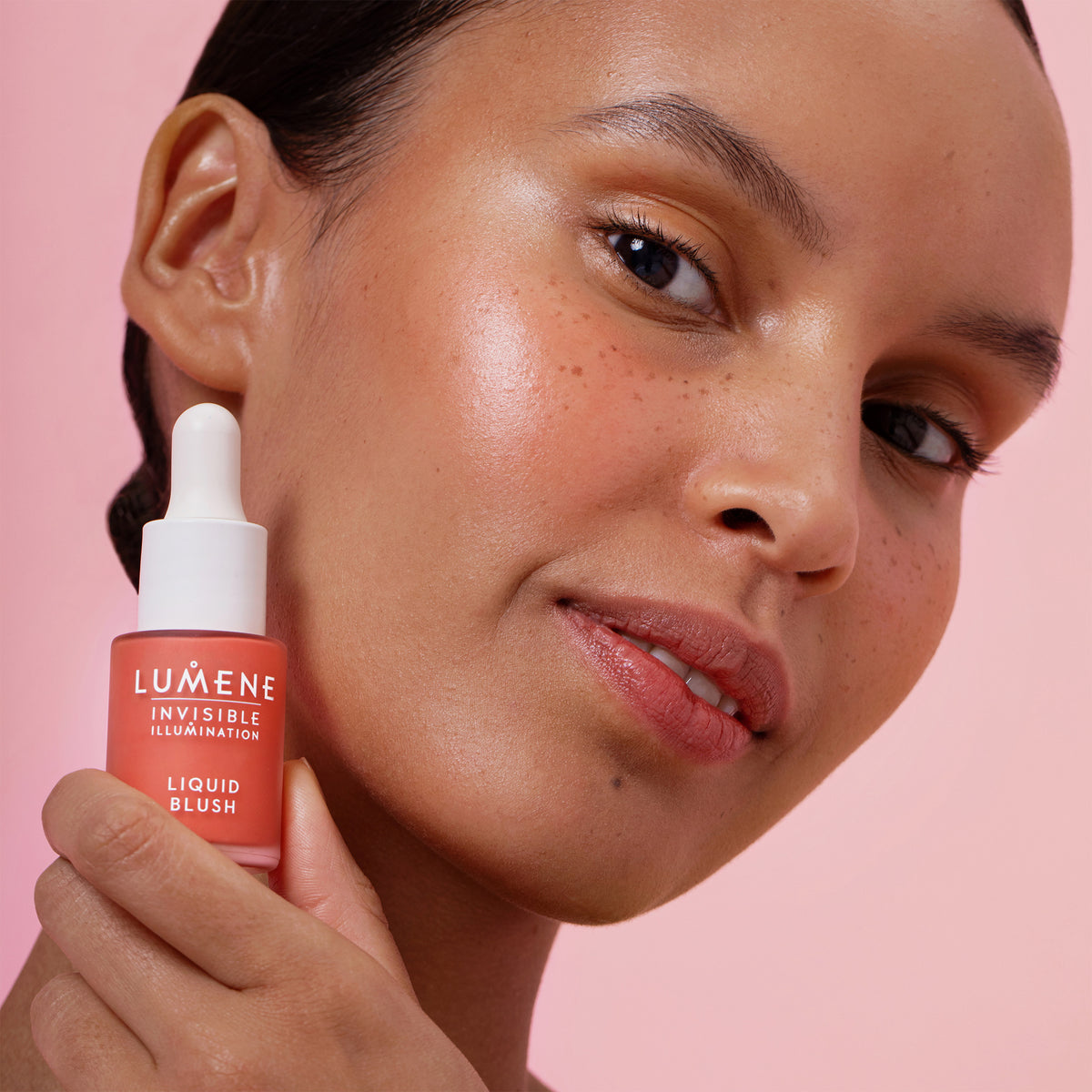 Eine Frau mit strahlendem mittlerem Hautton hält eine Flasche Lumene INVISIBLE ILLUMINATION Liquid Blush Bright Blossom an ihr Gesicht.