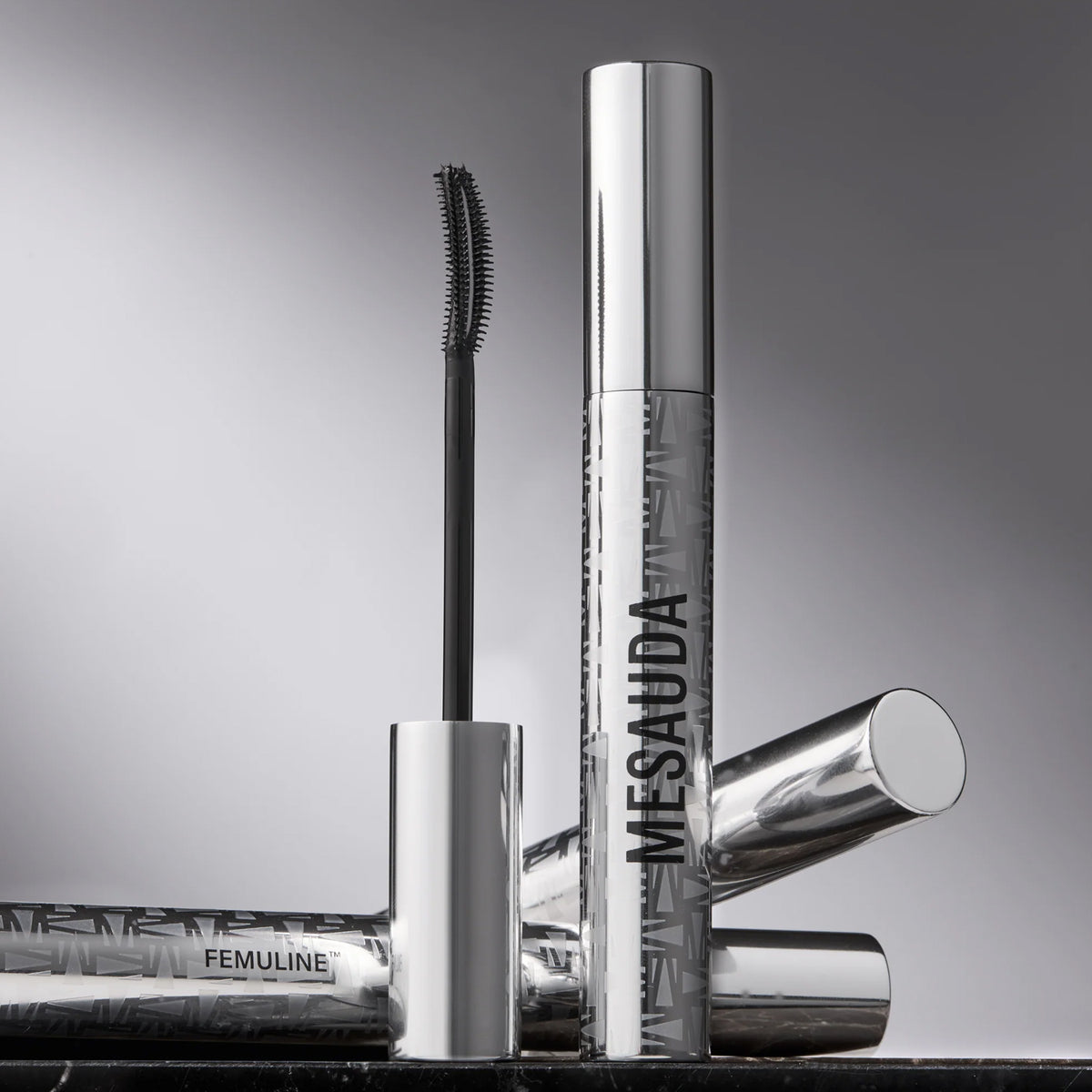 Eine silberne Tube Mesauda Beauty Femuline™ Mascara mit einer gebogenen Bürste wird auf einer reflektierenden Oberfläche präsentiert.