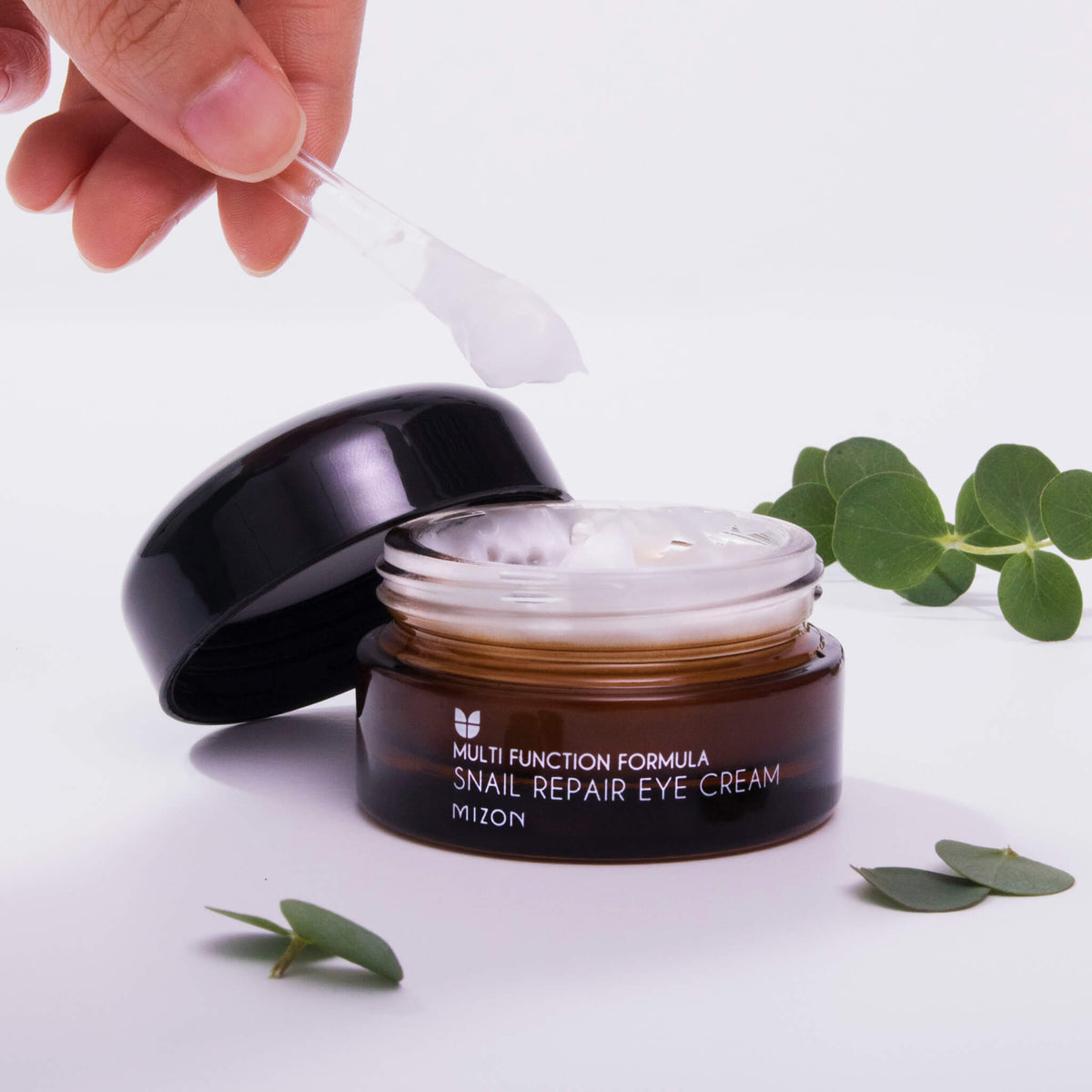 Eine Hand schöpft mit einem kleinen Spatel weiße Creme aus einem Tiegel mit Snail Repair Eye Cream.