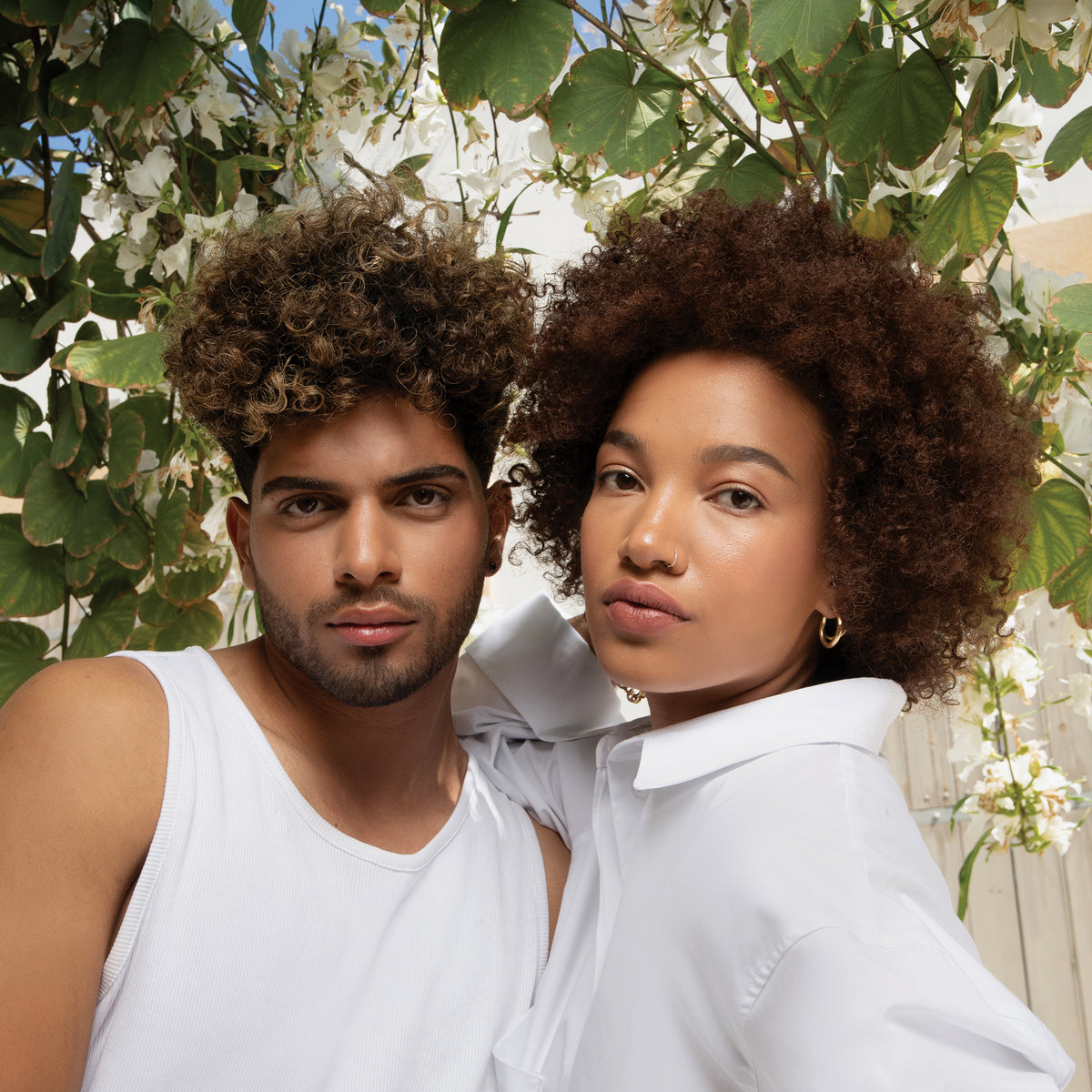 Zwei Menschen mit lockigem Haar stehen dicht beieinander im Freien, umgeben von grünen Blättern und weißen Blumen, und präsentieren Neumas NEU CURL™ PERFECTING CRÈME.