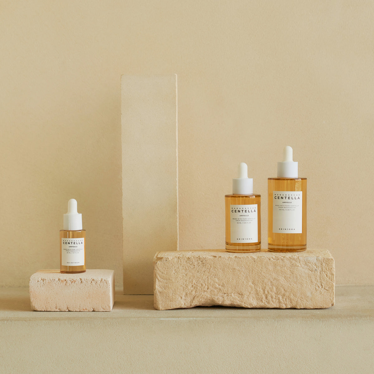 Drei Flaschen Madagascar Centella Ampoule sind auf beigen Steinblöcken vor einem neutralen Hintergrund ausgestellt.