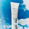 Eine Tube Madagascar Centella Hyalu-Cica Water-Fit Sun Serum steht aufrecht vor einem blauen Himmel mit Wolken und Wassertropfen.