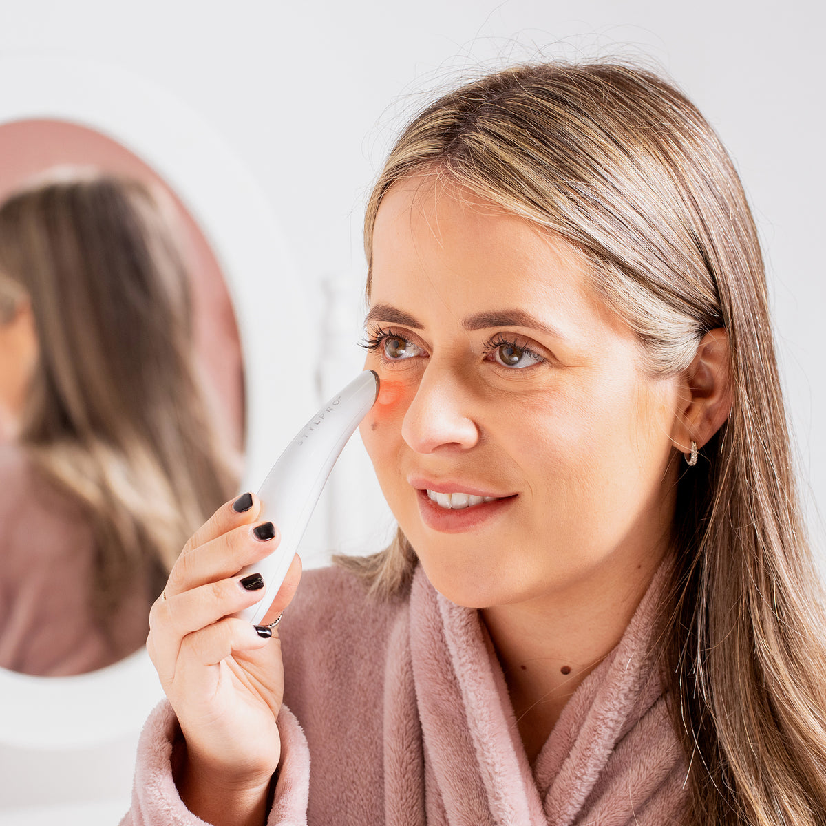 Eine Frau in einem rosa Bademantel verwendet vor einem Spiegel den Beauty Eye Massager unter ihrem Auge.