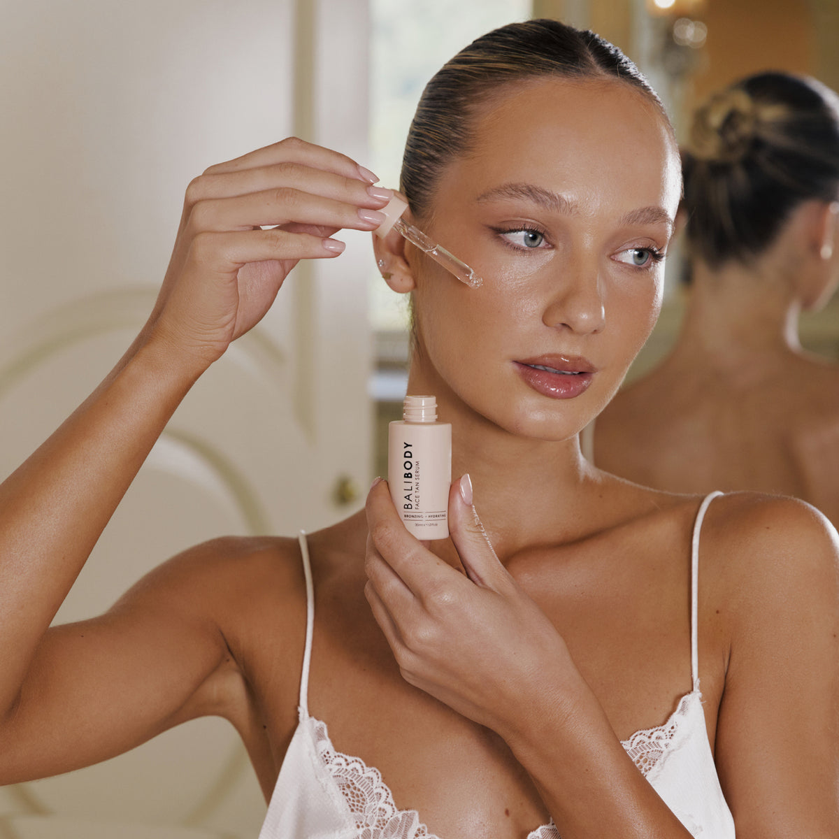 Eine Person in einem weißen Spitzentop steht in einem sanft beleuchteten Raum und trägt mit einer Pipette das Face Tan Serum auf