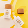 Zwei Flaschen DROP OF Sunshine BRONZING DROPS auf einem gelben Sonnenillustrationshintergrund.